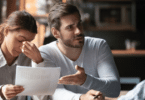 Um casal está em uma mesa. A mulher está com uma mão no rosto e tem a expressão de cansaço. Ao seu lado, o homem está reclamando com a pessoa na frente deles.