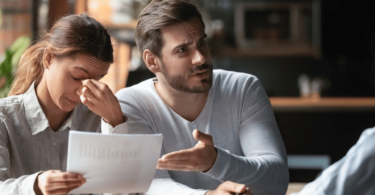 Um casal está em uma mesa. A mulher está com uma mão no rosto e tem a expressão de cansaço. Ao seu lado, o homem está reclamando com a pessoa na frente deles.