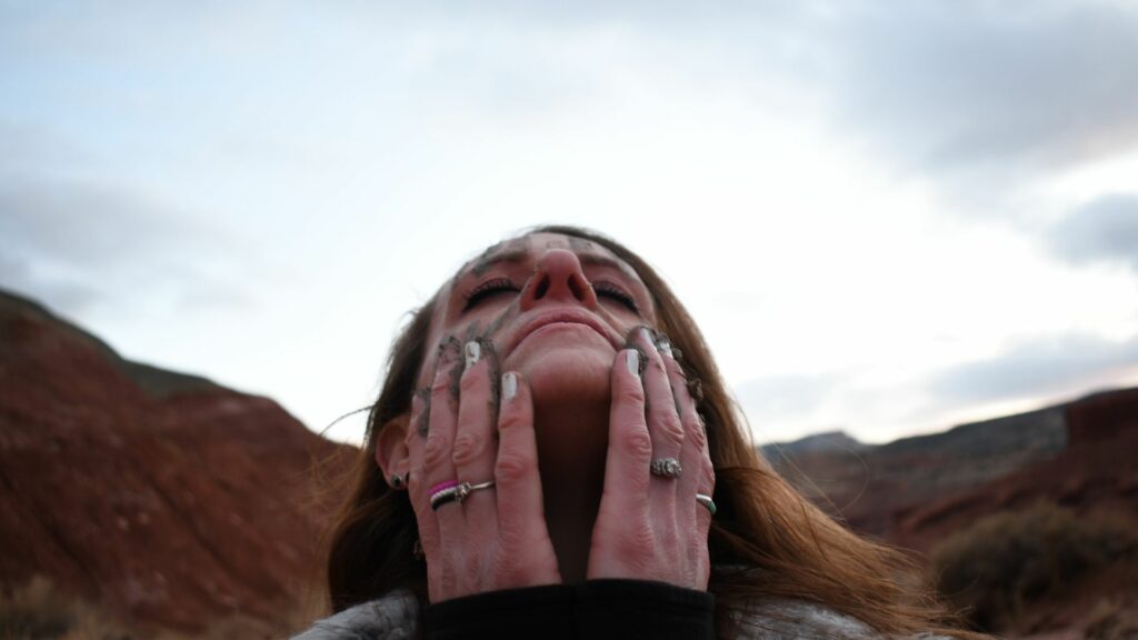 Imagem de fundo de uma montanha e em destaque uma mulher olhando para o céu de olhos fechados e com as duas mãos sobre o rosto, sujas de terra, trazendo o conceito de renascimento pessoal.
