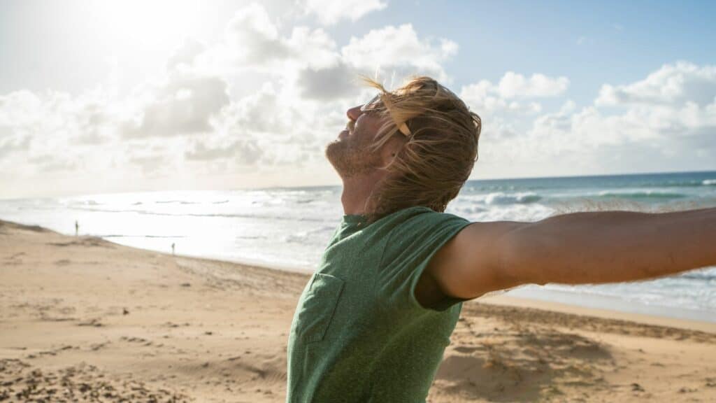 Imagem de uma praia em um dia de sol. Em destaque, um homem alegre, feliz de braços abertos, se aceitando como é.