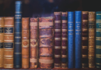 Quinze livros antigos estão organizados lado a lado em uma prateleira de madeira.