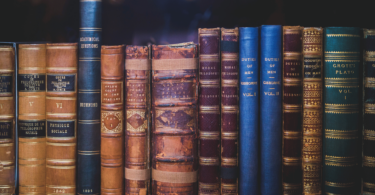 Quinze livros antigos estão organizados lado a lado em uma prateleira de madeira.