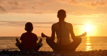 Mãe e filho estão sentados de costas para a imagem e meditam de frente para o mar e para o pôr do sol.