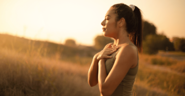 Uma mulher jovem está ao ar livre. Ela respira o ar fresco, está com os olhos fechados, junta as duas mãos e as coloca sobre o peito, como se estivesse meditando na natureza.