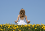 Uma mulher está sentada de costas para a imagem ao ar livre. Ela faz a posição da meditação e observa um céu azul claro. Ela está sentada em um campo cheio de flores amarelas.