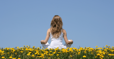 Uma mulher está sentada de costas para a imagem ao ar livre. Ela faz a posição da meditação e observa um céu azul claro. Ela está sentada em um campo cheio de flores amarelas.