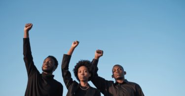 Três pessoas negras, uma mulher e dois homens, estão no foco da imagem. Eles levantam os seus punhos das mãos direitas no ar. Eles usam roupas pretas. Ao fundo, há um céu azul claro.