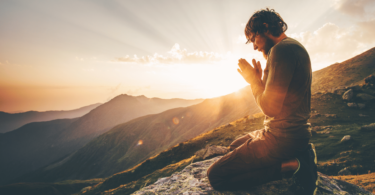 Um homem ajoelhado com as duas mãos unidas em sinal de oração está no foco da imagem. Ele está com os olhos fechados e a cabeça inclinada para baixo. Ele está em um ambiente de natureza, em cima de uma montanha. Ao fundo, há montanhas e o pôr do sol.