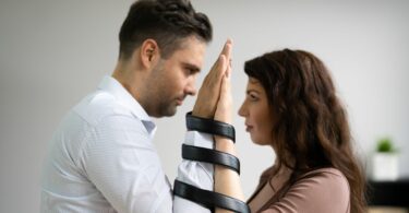 Imagem de um casal olhando um para o outro de mãos levantadas, tendo os braços entrelados por uma fita preta, trazendo o conceito de uma relação tóxica.