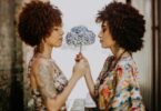 Imagem de duas mulheres frente a frente segurando uma flor de pétalas brancas e azuis, trazendo o conceito de compartilhar.