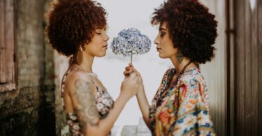 Imagem de duas mulheres frente a frente segurando uma flor de pétalas brancas e azuis, trazendo o conceito de compartilhar.