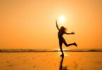 Imagem do pôr do sol em uma praia e em destaque a silhueta de uma moça feliz e radiante, trazendo o conceito de uma pessoa iluminada.