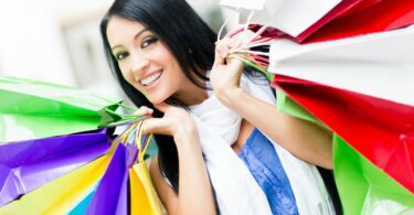 Imagem de uma jovem mulher, com cabelos longos e preto, segurando várias sacolas coloridas, trazendo o conceito de compras por compulsão.