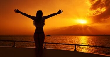 Imagem de uma mulher de braços abertos, olhando para o pôr do Sol, trazendo o conceito de alma limpa e gratidão.