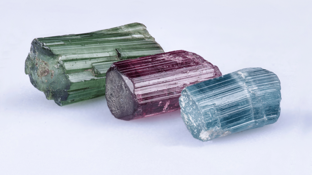 Três pedras turmalinas de cores distintas (verde, rosa e azul) estão no foco da imagem.