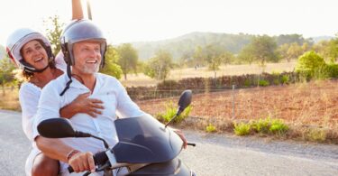 Imagem de um casal maduro passeando de moto em uma estrada cheia de montanhas e áreas verdes.