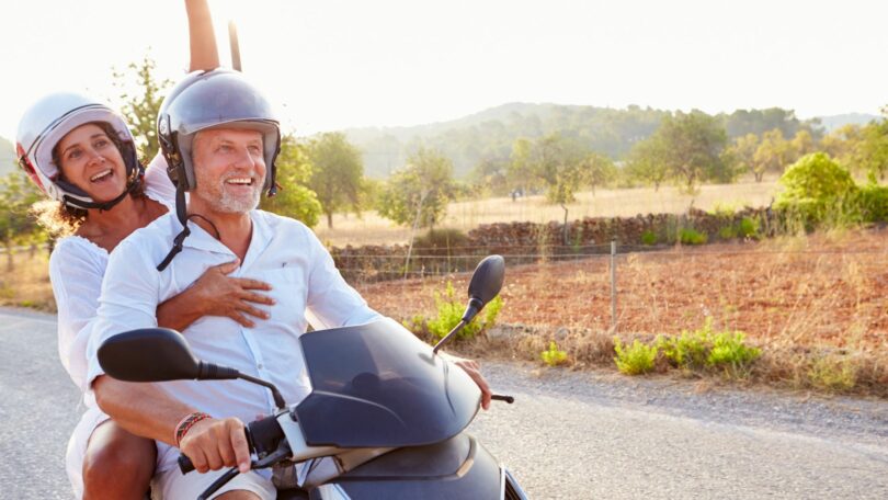 Imagem de um casal maduro passeando de moto em uma estrada cheia de montanhas e áreas verdes.
