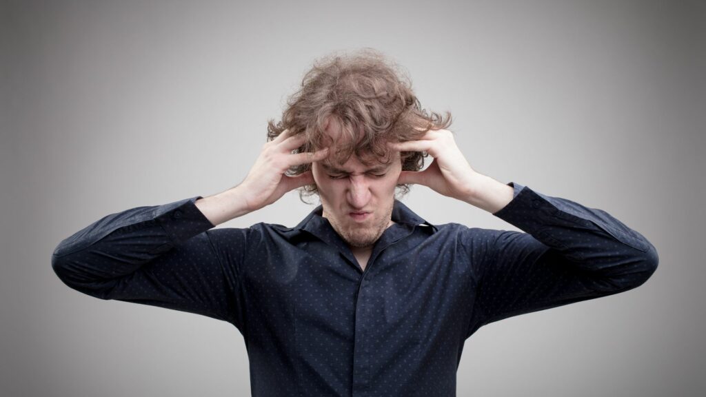 Imagem de um homem usando uma camisa social azul marinho. Ele está com as mãos na cabeça. A imagem traz o conceito sobre pensar demais, algo que pode ser estressante e perigoso.
