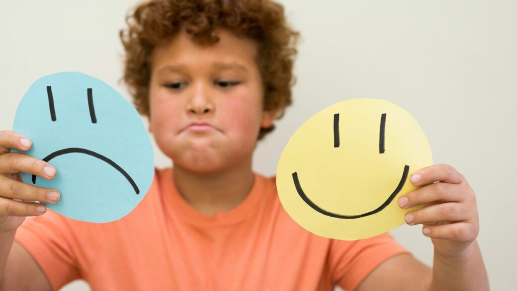 Imagem de uma criança segurando em suas mãos emojis com sentimentos e emoções de alegria e tristeza.
