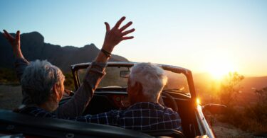 Imagem de um casal 60+ dentro de um carro sem teto, olhando o pôr do sol. Eles estão felizes, vivendo o momento presente.