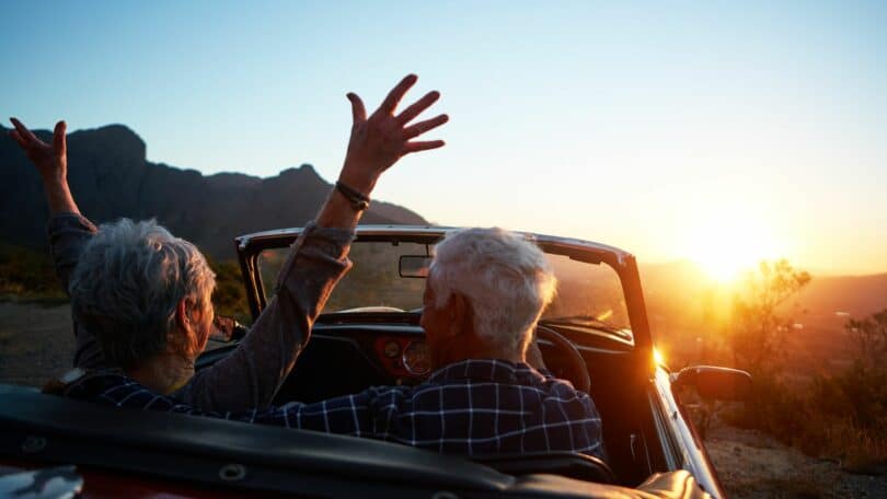 Imagem de um casal 60+ dentro de um carro sem teto, olhando o pôr do sol. Eles estão felizes, vivendo o momento presente.
