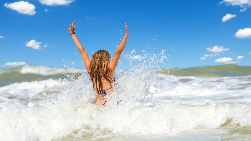 Imagem de uma jovem de cabelos longos dourados dentro do mar, com as mãos levantadas, curtindo o presente momento, vivendo a vida com paixão.
