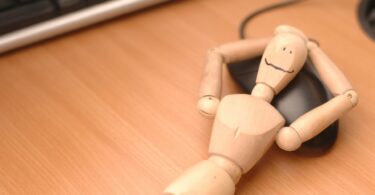 Imagem de um boneco de madeira deitado sobre um mouse, de barriga para cima em uma mesa de computador, trazendo o conceito de pausa das redes digitais.