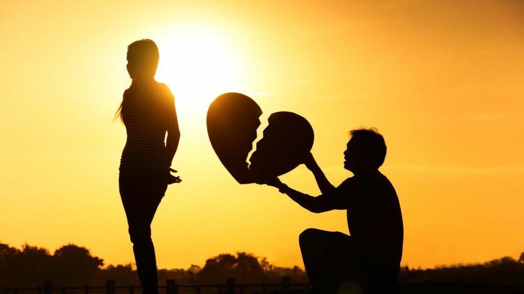 Imagem de um lindo pôr do sol e em destaque a silhueta de um casal. Ele está segurando um coração partido ao meio, simbolizando o fim do amor.