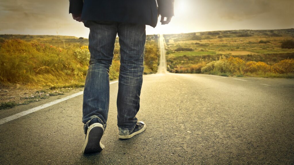 Imagem de um homem usando jeans, um casaco preto e um tênis, caminhando humildemente em uma estrada deserta.
