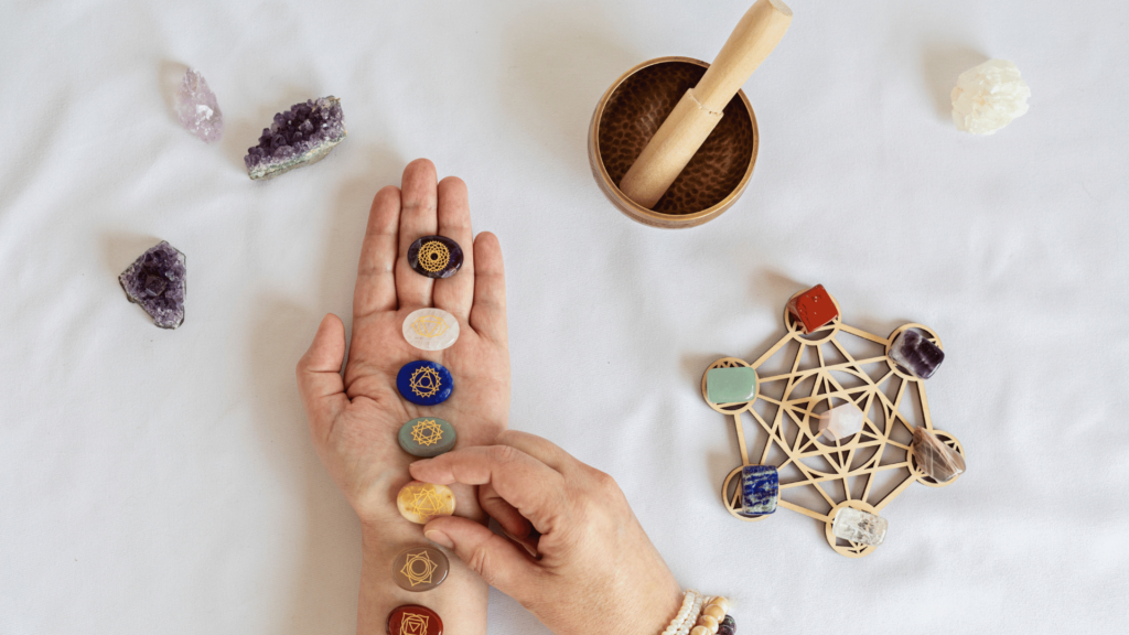 As mãos de uma pessoa estão no foco da imagem. Ela coloca uma fileira de pedras sobre sua mão esquerda. E, sobre a mesa, há outras pedras e objetos de meditação.