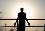 A silhueta de um homem está ao ar livre na frente de uma cerca. Ele observa o ambiente de natureza. A luz do sol reflete forte ao fundo.