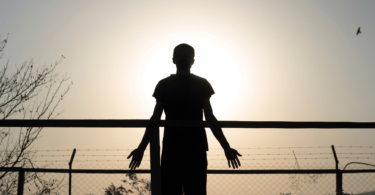 A silhueta de um homem está ao ar livre na frente de uma cerca. Ele observa o ambiente de natureza. A luz do sol reflete forte ao fundo.