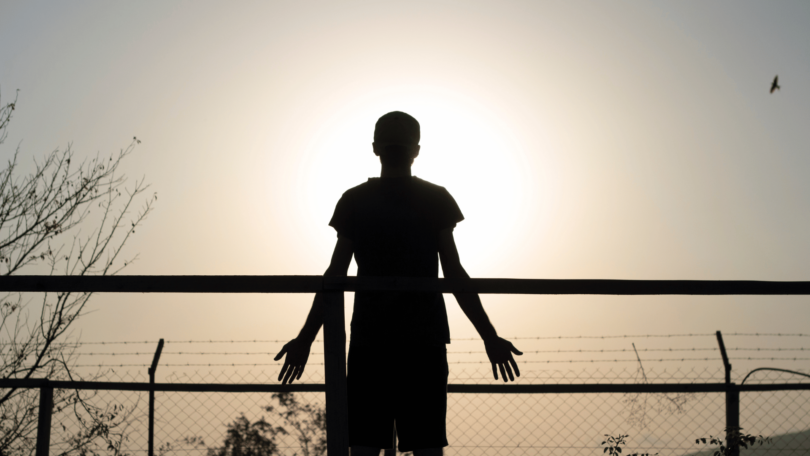 A silhueta de um homem está ao ar livre na frente de uma cerca. Ele observa o ambiente de natureza. A luz do sol reflete forte ao fundo.