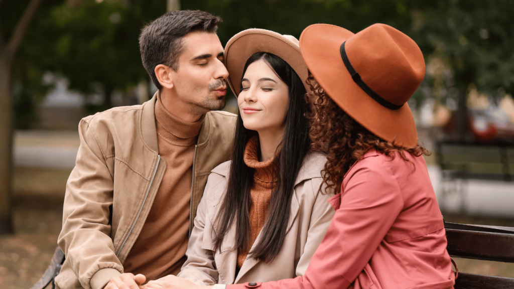 Um homem e duas mulheres estão sentados em um banco de um parque, representando o conceito de poliamor.