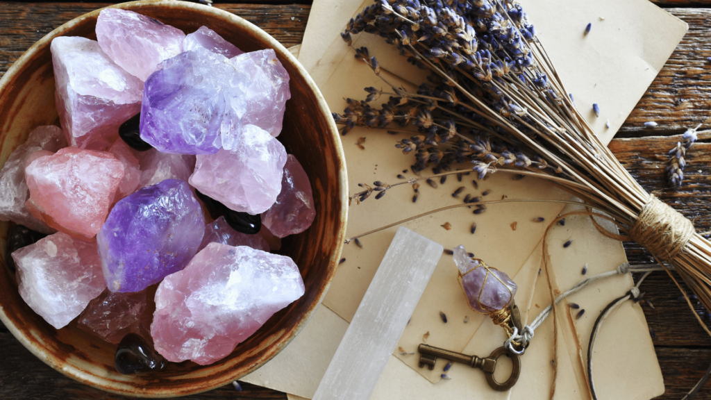 No foco da imagem há um recipiente com vários cristais de quartzo rosa. Ao lado, há alguns papéis antigos e uma lavanda seca.