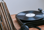 No foco da imagem há um disco de vinil tocando em um aparelho "toca-discos". E ao lado dele, há algumas capas de discos de vinil empilhadas e um fone de ouvido.