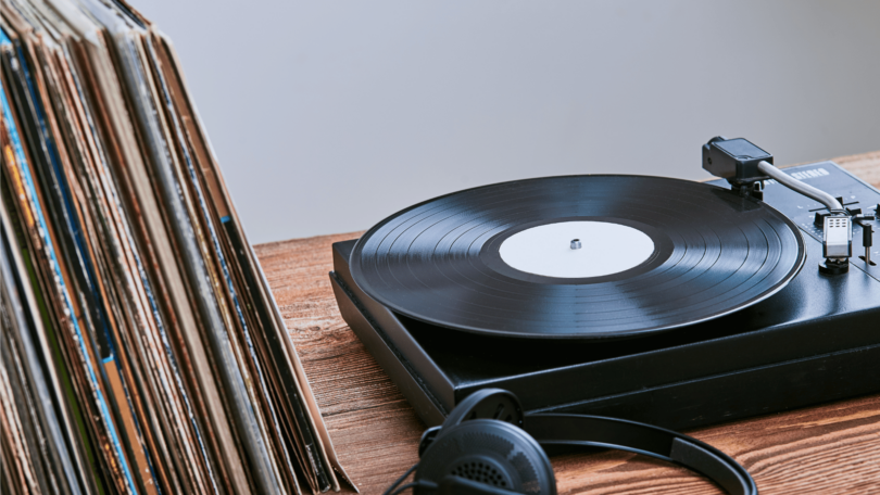 No foco da imagem há um disco de vinil tocando em um aparelho "toca-discos". E ao lado dele, há algumas capas de discos de vinil empilhadas e um fone de ouvido.