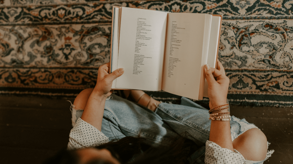 Uma mulher segurando um livro de poesia aberto está no centro da imagem. Ela está sentada no chão lendo o livro.