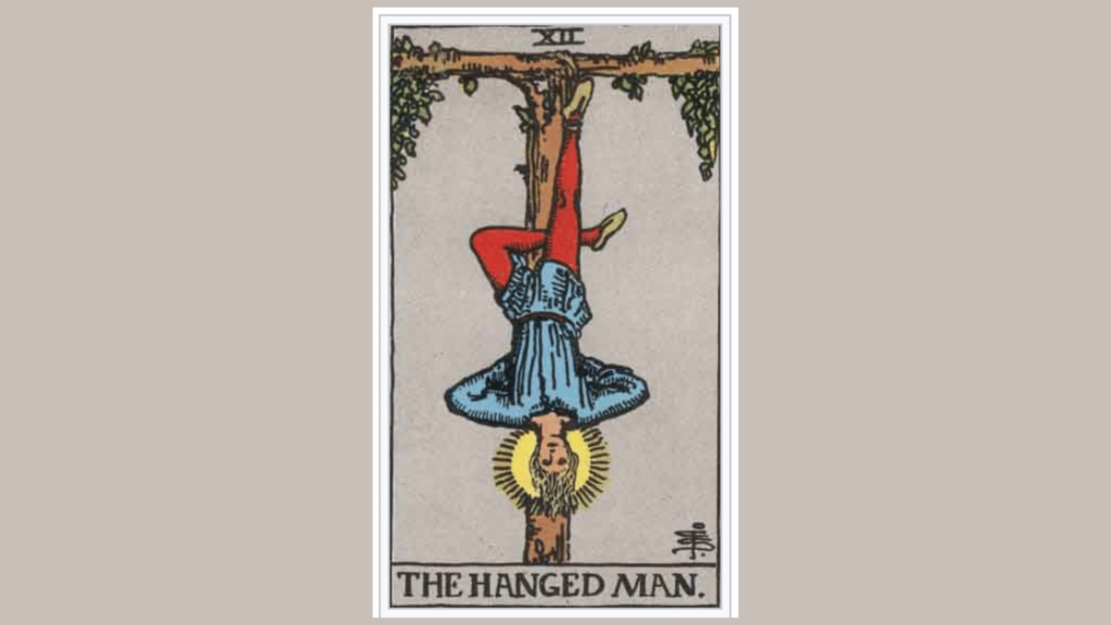 A carta do Tarot "O Enforcado" está no centro da imagem. Há um fundo cinza.