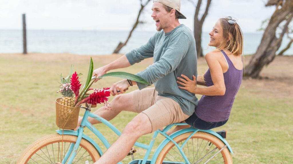 Imagem de um casal andando de bicicleta na beira da praia, trazendo o conceito do quinto princípio Huna, o Aloha, que significa "olá", "adeus" e também tem um significado mais profundo como amor, compaixão, respeito e conexão.
