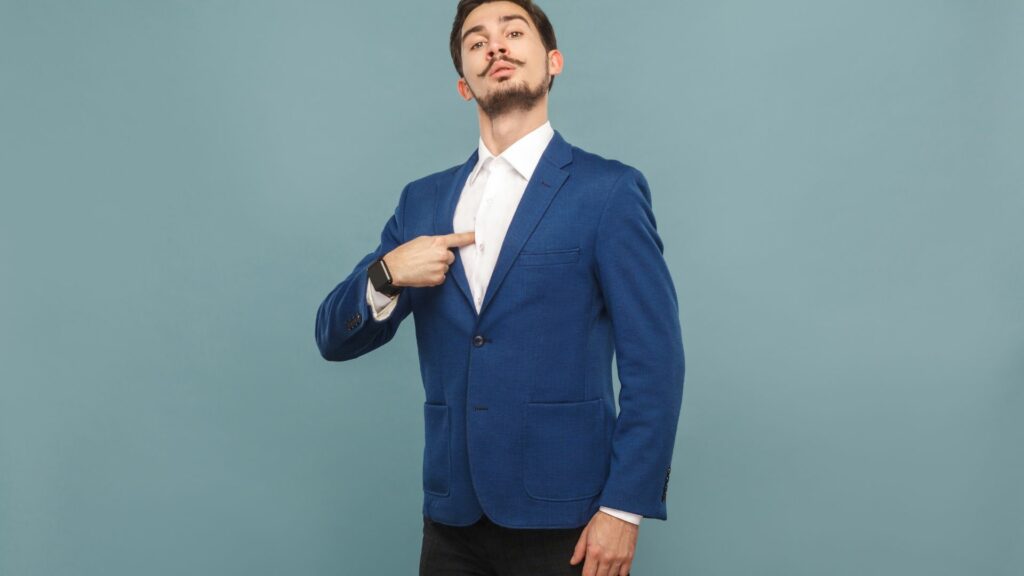 Imagem de um homem bem vestido, usando o terno azul e uma camisa branca. Ele está apontando o dedo para si mesmo, se achando um homem de sucesso. A imagem traz o conceito de ego.
