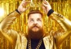 Imagem de fundo dourado. Em destaque um homem de cabelo e barba ruivo, usando um blazer e uma corrente dourada. Ele está colocando sobre a sua cabeça uma coroa, também dourada, simbolizando a supremacia do ego.
