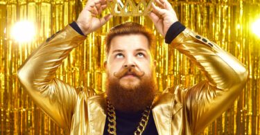 Imagem de fundo dourado. Em destaque um homem de cabelo e barba ruivo, usando um blazer e uma corrente dourada. Ele está colocando sobre a sua cabeça uma coroa, também dourada, simbolizando a supremacia do ego.