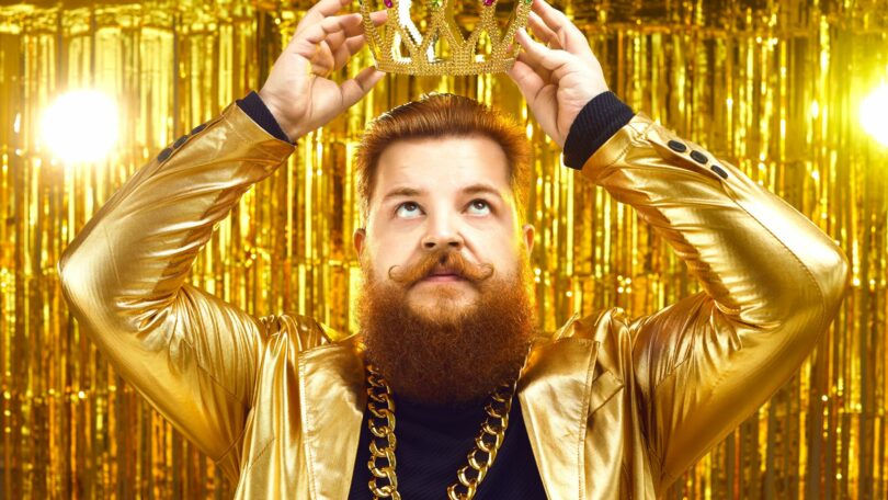 Imagem de fundo dourado. Em destaque um homem de cabelo e barba ruivo, usando um blazer e uma corrente dourada. Ele está colocando sobre a sua cabeça uma coroa, também dourada, simbolizando a supremacia do ego.