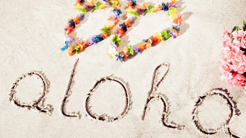 Imagem da frase ALOHA escrita na areia de uma praia, ao lado arranjos de flores, trazendo o conceito de amor e respeito.