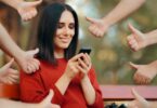 Imagem de uma mulher olhando o celular e as suas redes sociais. Ela está rodeada de mãos fazendo o sinal de jóia, ou seja, está sendo aprovada por suas postagens nas redes sociais.