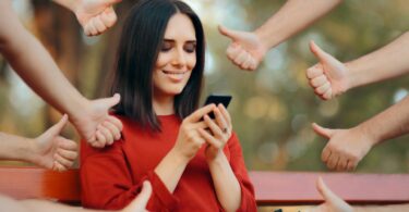 Imagem de uma mulher olhando o celular e as suas redes sociais. Ela está rodeada de mãos fazendo o sinal de jóia, ou seja, está sendo aprovada por suas postagens nas redes sociais.