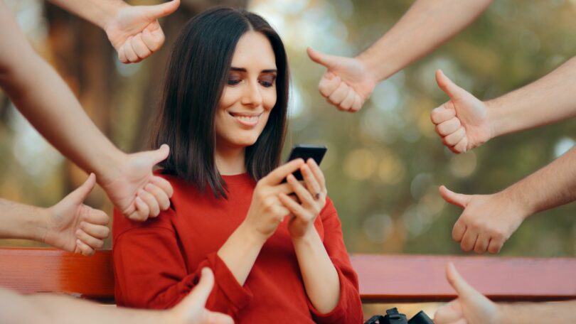 Imagem de uma mulher olhando o celular e as suas redes sociais. Ela está rodeada de mãos fazendo o sinal de jóia, ou seja, está sendo aprovada por suas postagens nas redes sociais.