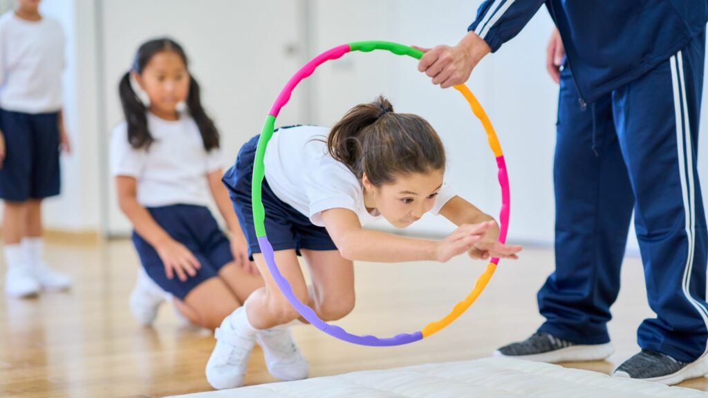 Imagem de várias crianças em uma sala de aula fazendo exercícios. Em destaque, uma delas realizando um salto de arco na aula de ginástica com o seu professor.
