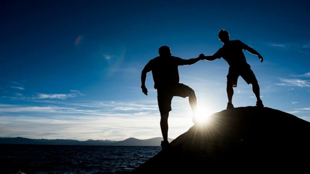 Imagem de um pôr do Sol e em destaque a silhueta de dois amigos sobre uma pedra, de mãos dadas, trazendo o conceito de ajuda mútua.
